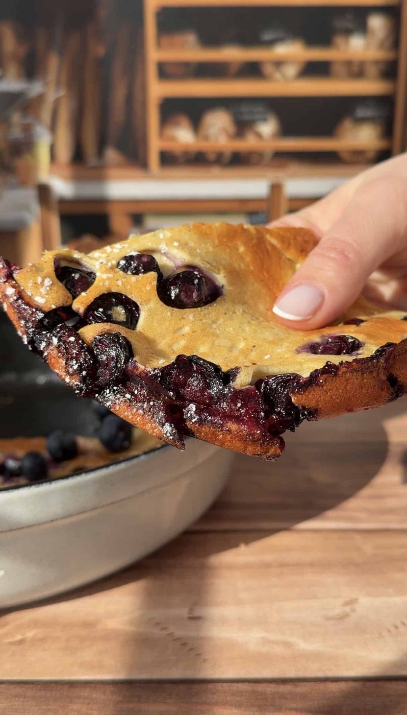 A hand holds a thick, golden-brown slice of High Protein Dutch Baby pancake, with juicy blueberries visible throughout. A ceramic dish with more pancake is in the background on a wooden table.