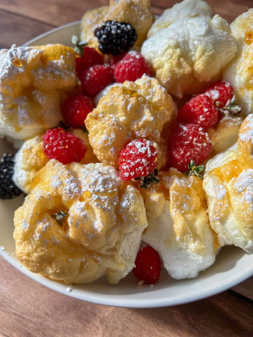 A bowl of fluffy cream puffs dusted with powdered sugar, garnished with fresh raspberries and blackberries, sits beside High Protein Mini Pancakes on a rustic wooden table.
