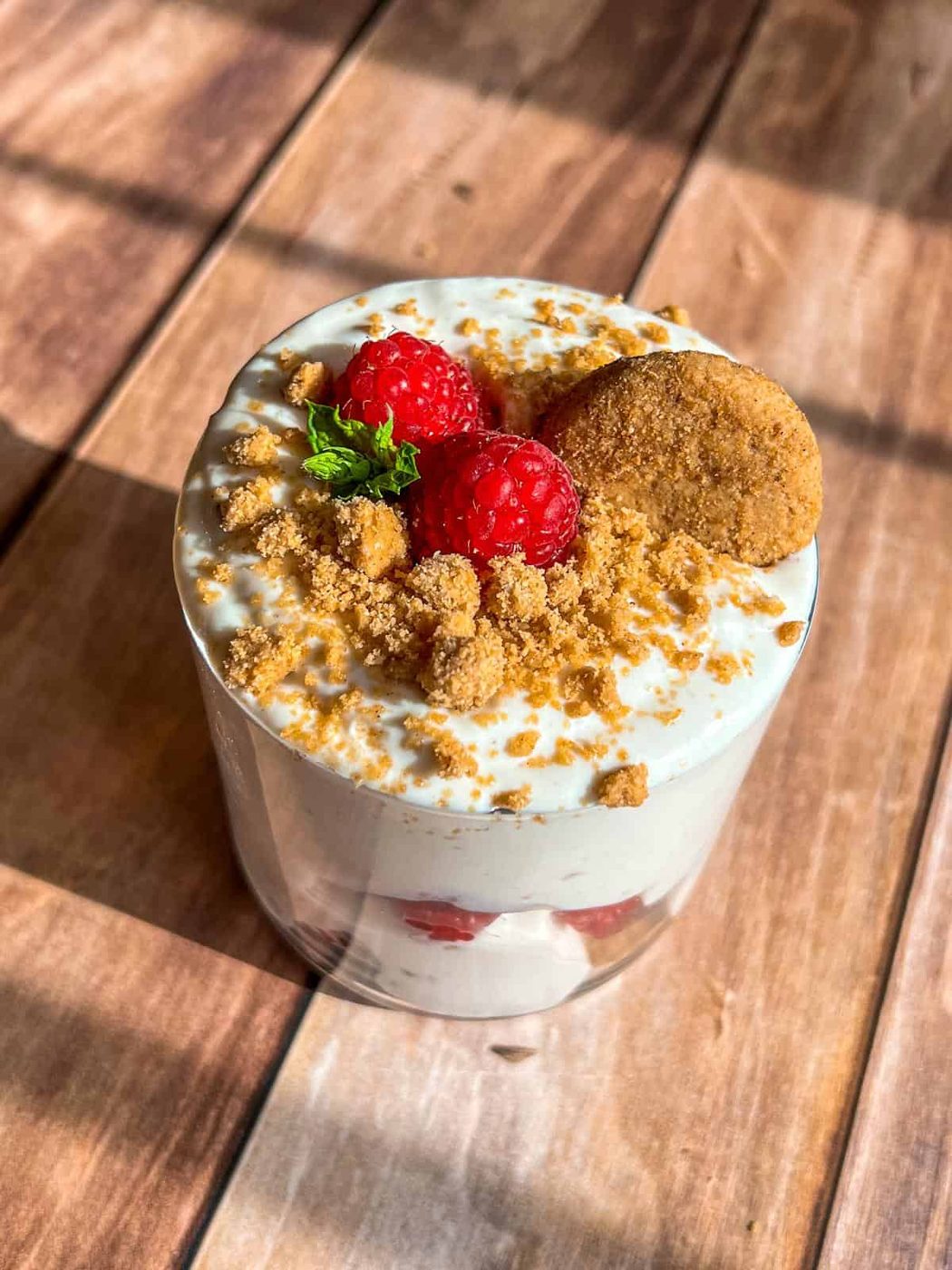 High Protein Pudding. A dessert cup with creamy layers, topped with fresh raspberries, crumbled cookie, a whole cookie, and a small mint leaf, placed on a wooden surface with sunlight casting shadows.