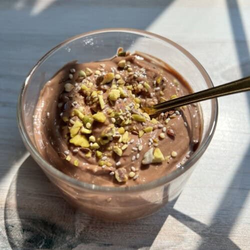 A clear glass filled with creamy chocolate pudding, topped with chopped nuts and seeds, sits on a sunlit wooden surface. A gold spoon is placed inside the glass. Shadows from window panes fall across the scene.