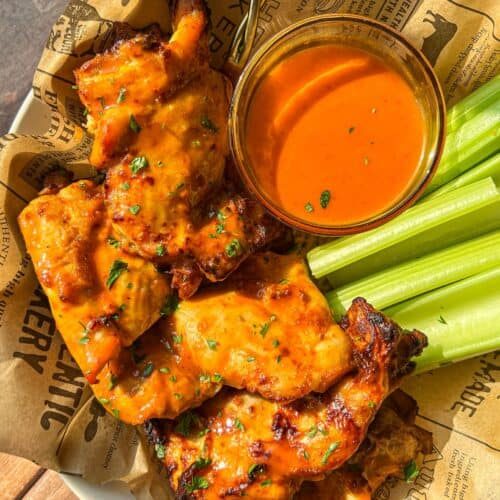 A plate of saucy grilled chicken wings with fresh celery sticks and a bowl of orange dipping sauce, served on printed parchment paper.