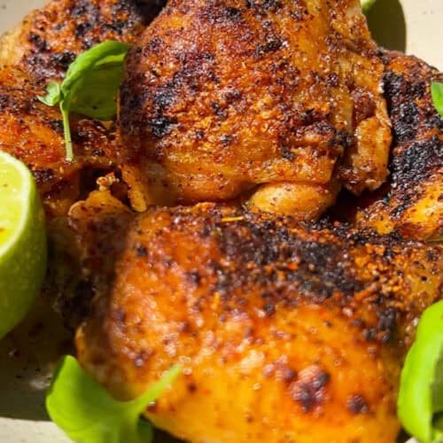 Close-up of a dish featuring well-seasoned, grilled chicken thighs with charred skin, garnished with fresh green leaves. A halved lime is placed beside the chicken, adding a zesty touch to the presentation.