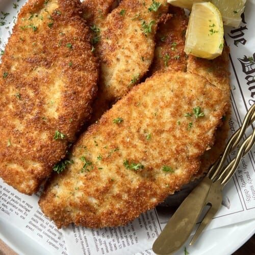 Three golden-brown breaded schnitzels on a white plate, garnished with chopped parsley and served with lemon wedges. A fork and knife rest on the plate, which is lined with printed paper.