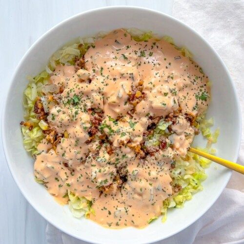 A white bowl filled with chopped lettuce, ground beef, and a generous layer of creamy orange Big Mac Salad sauce, garnished with herbs. A yellow fork rests in the bowl, which sits on a white surface next to a light napkin.
