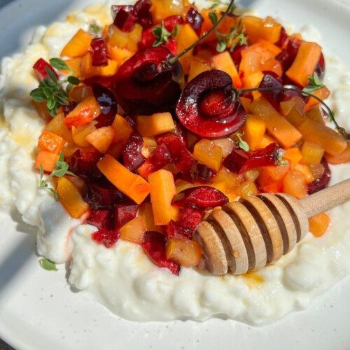 A plate of creamy Cottage Cheese Fruit Salad topped with chopped cherries, apricots, fresh herbs, and a honey dipper drizzled with honey, served on a white dish in bright natural light.