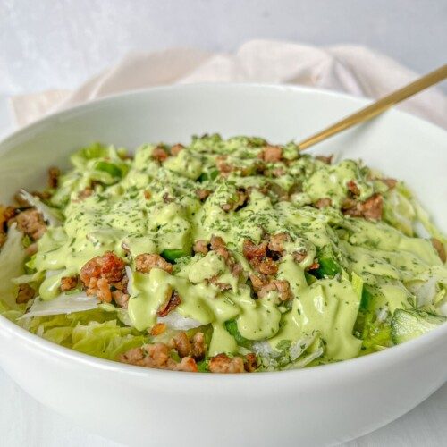 A white bowl filled with a green salad topped with crumbled meat and drizzled with a creamy green dressing—perfect for using fresh greens after learning how to dry herbs—rests beside a fork ready for your first bite.