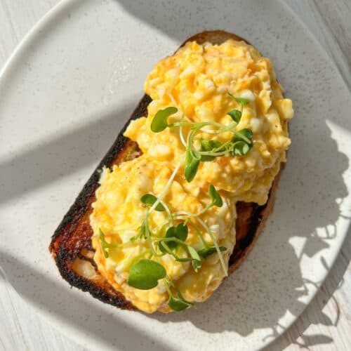 A plate with two slices of toast topped with scrambled eggs and garnished with microgreens. The toast is slightly charred, and sunlight casts shadows on the white, speckled plate and wooden table surface.