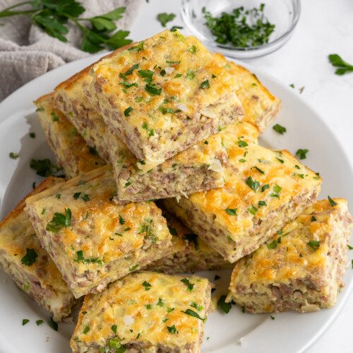 A plate of square-shaped sausage and cheese breakfast casserole pieces, garnished with chopped parsley. A small glass bowl with chopped herbs and a beige napkin are in the background.