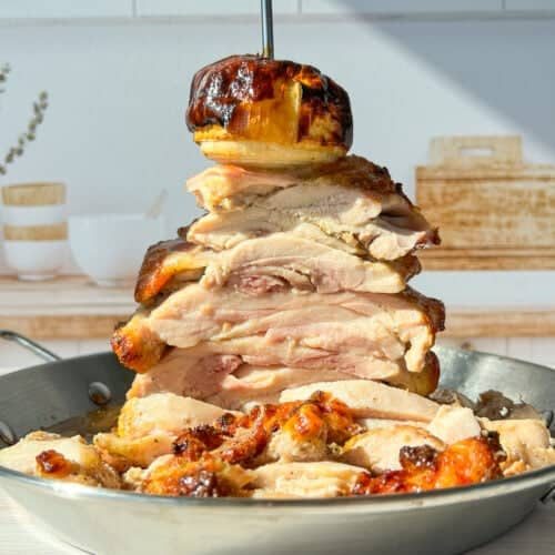 A towering stack of sliced, roasted chicken pieces is skewered on a vertical rod in a pan. A roasted onion tops the stack. The setting includes a bright kitchen with a wooden cutting board and decorative jars in the background.