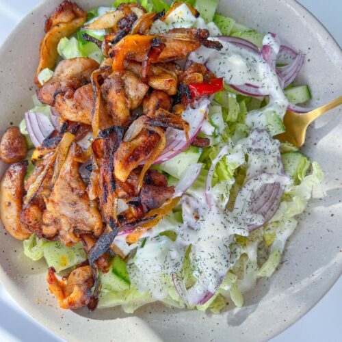 A bowl of Chicken Fajita Salad topped with grilled chicken, sliced red onions, lettuce, cucumber, and drizzled with a creamy dressing, served with a gold fork.