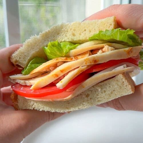 A close-up of a hand holding a sandwich with sliced turkey, tomato, lettuce, and cheese between two thick slices of bread, with a blurred window in the background.