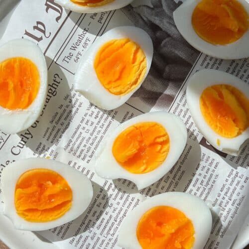 Several hard-boiled eggs, sliced in half to reveal their bright orange yolks, are arranged on a white plate lined with a sheet of newspaper. Sunlight casts shadows across the eggs and newspaper.