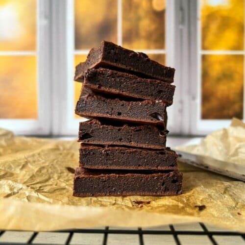 A stack of six chocolate brownies is displayed on parchment paper. The background features a blurred view of a window with autumn foliage visible outside. The scene is warmly lit, creating a cozy atmosphere.