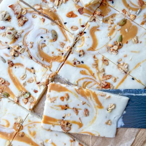 A close-up of Cottage Cheese Bark dessert bars with a creamy white topping swirled with caramel and sprinkled with granola, sitting on parchment paper. A knife is partially placed under one of the bars.