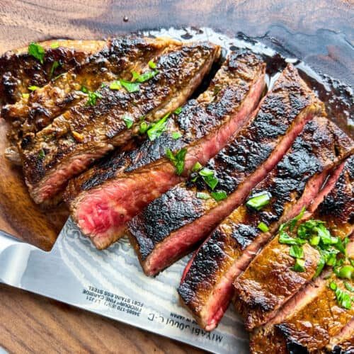 Sliced medium-rare steak with a seared crust, inspired by a classic Carne Asada recipe, garnished with chopped herbs and displayed on a wooden cutting board with a knife partially visible underneath.