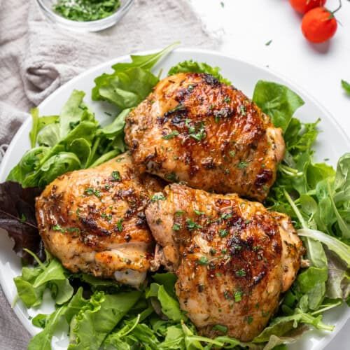 Grilled chicken thighs garnished with herbs are served on a bed of mixed greens on a white plate. A small bowl of chopped herbs and a bunch of fresh cherry tomatoes are in the background.