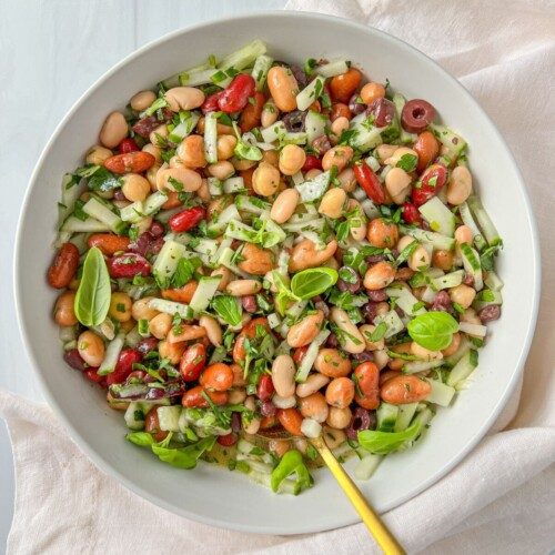 A white bowl filled with a vibrant Mixed Bean Salad of chopped cucumber, red onion, corn, fresh herbs, and basil leaves rests on a light cloth with a golden fork inside the bowl.