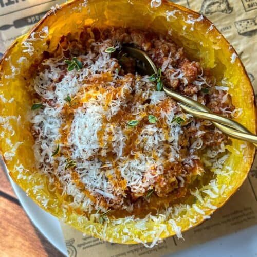 A baked spaghetti squash filled with tomato meat sauce and topped with grated cheese, served in its shell with a spoon. The dish sits on a white plate and a sheet of brown paper.