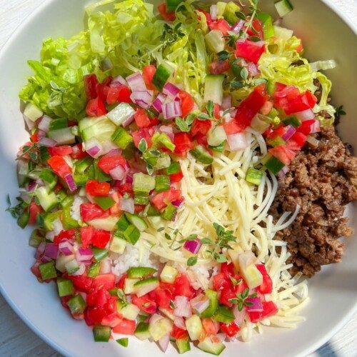 A Beef Burrito Bowl Recipe comes to life with shredded lettuce, diced tomatoes, cucumbers, red onions, cooked ground beef, and spaghetti noodles, all artfully arranged in sections on a white surface with sunlight streaming in.