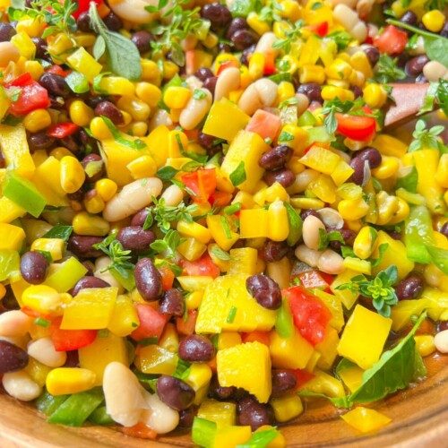 A close-up of Cowboy Caviar in a wooden bowl, featuring black beans, white beans, corn, diced red and yellow bell peppers, tomatoes, and fresh green herbs.