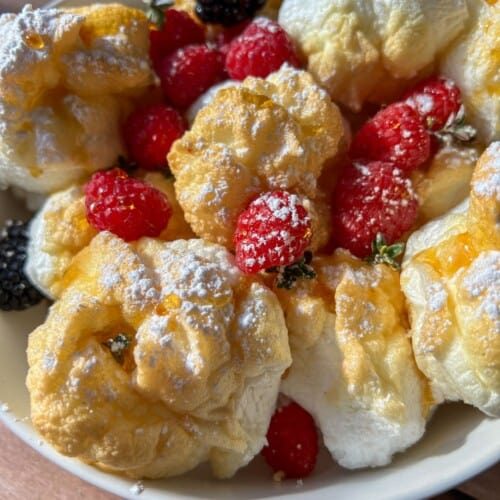 A bowl of fluffy cream puffs dusted with powdered sugar, garnished with fresh raspberries and blackberries, sits beside High Protein Mini Pancakes (Viral) on a rustic wooden table.
