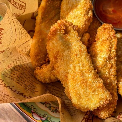 Golden, crispy Healthy Chicken Tenders served in a basket lined with printed paper, with a small metal cup of red dipping sauce on the side.