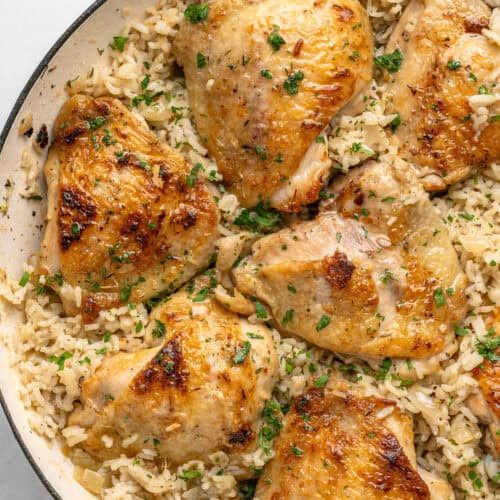 Golden-brown chicken thighs on a bed of seasoned rice, garnished with chopped parsley and inspired by the bold flavors of Mexican Hot Dogs, served in a white skillet.