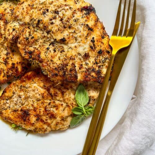 Grilled, seasoned chicken breasts on a white plate garnished with fresh basil, next to a gold fork and knife, set on a white cloth background.