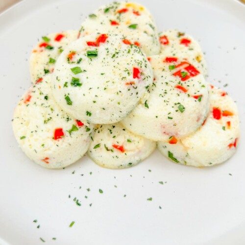 A plate with a stack of round, fluffy Copycat Starbucks Egg-Bites with Ricotta, speckled with red bell peppers and green herbs, presented on a white plate and lightly garnished with chopped herbs.