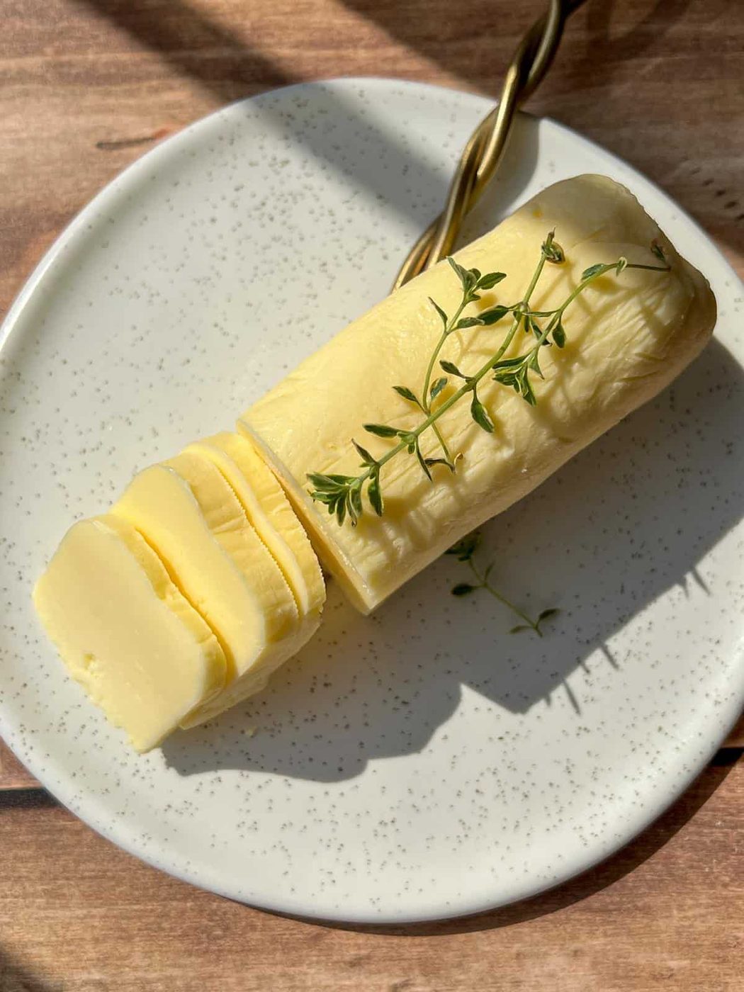 A log of sliced butter garnished with fresh thyme sits on a speckled white plate, with a gold-handled knife beside it, all on a wooden surface lit by sunlight.