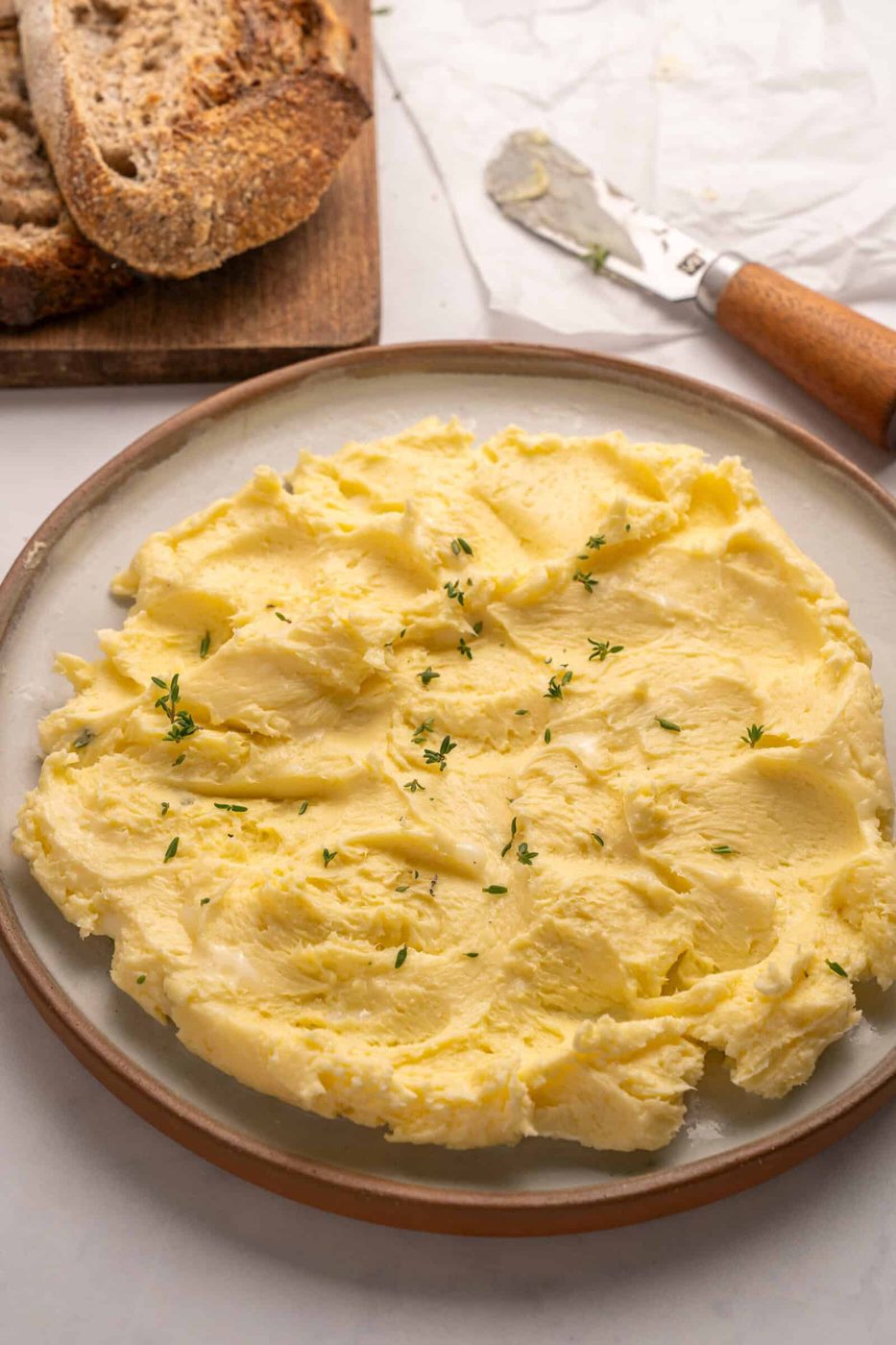 A plate of creamy, whipped butter garnished with fresh herbs sits on a table next to slices of rustic bread and a butter knife.
