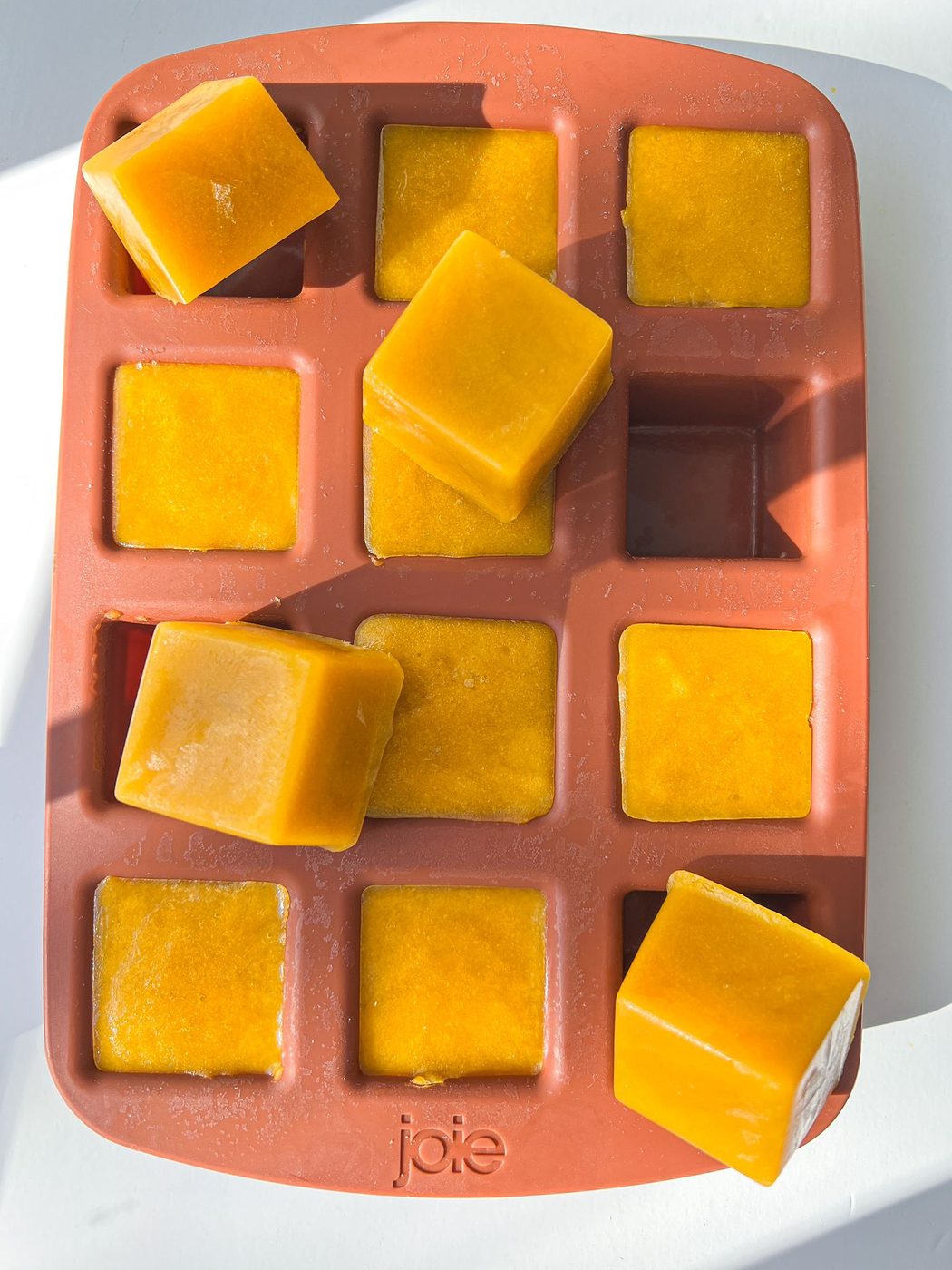 A red silicone ice cube tray holds several orange-yellow Homemade Chicken Bouillon Cubes, some neatly inside the tray and others resting on top, all in bright sunlight.
