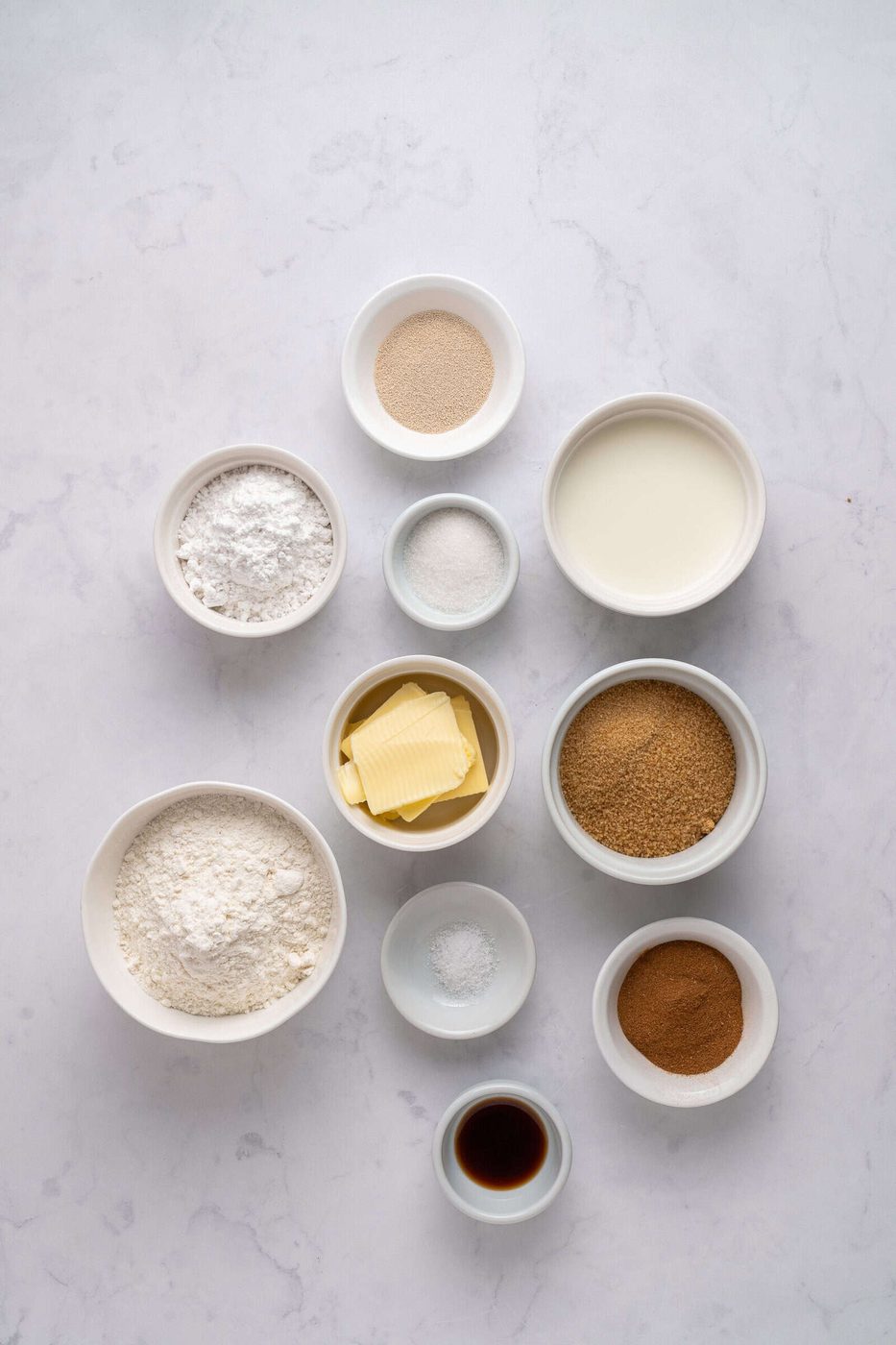 An overhead view of ten small bowls containing baking ingredients, including flour, yeast, sugar, milk, butter slices, brown sugar, cinnamon, vanilla extract, and salt, arranged neatly on a light marble surface.