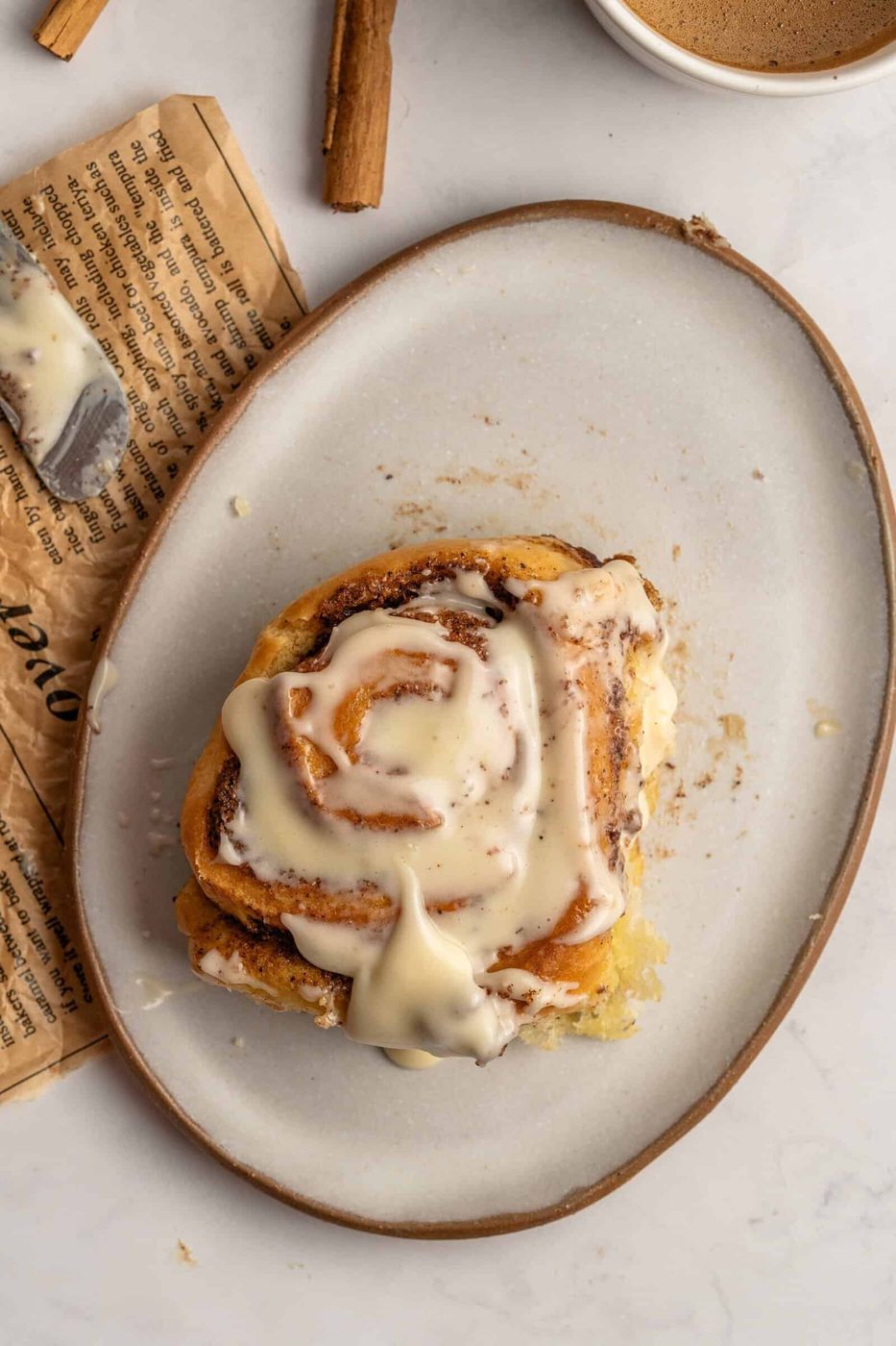 A close-up of a cinnamon roll from a Homemade Cinnamon Rolls topped with creamy icing sits on an oval plate, with part of a newspaper and a cinnamon stick nearby on a marble surface.