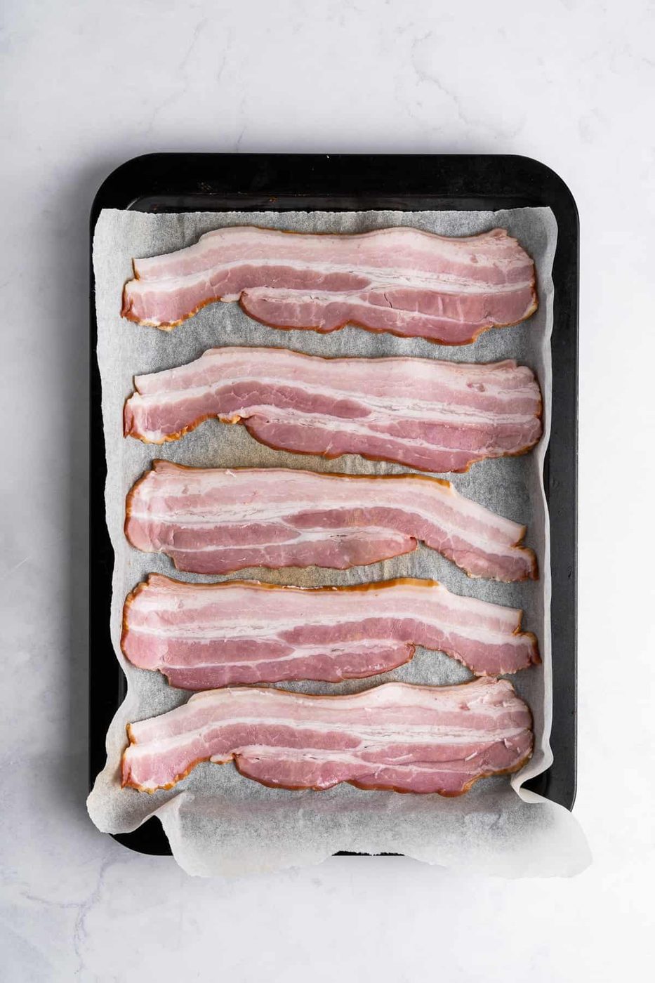 Five uncooked bacon strips are arranged neatly on a parchment-lined baking tray. The tray rests on a light-colored marble surface, ready for cooking.