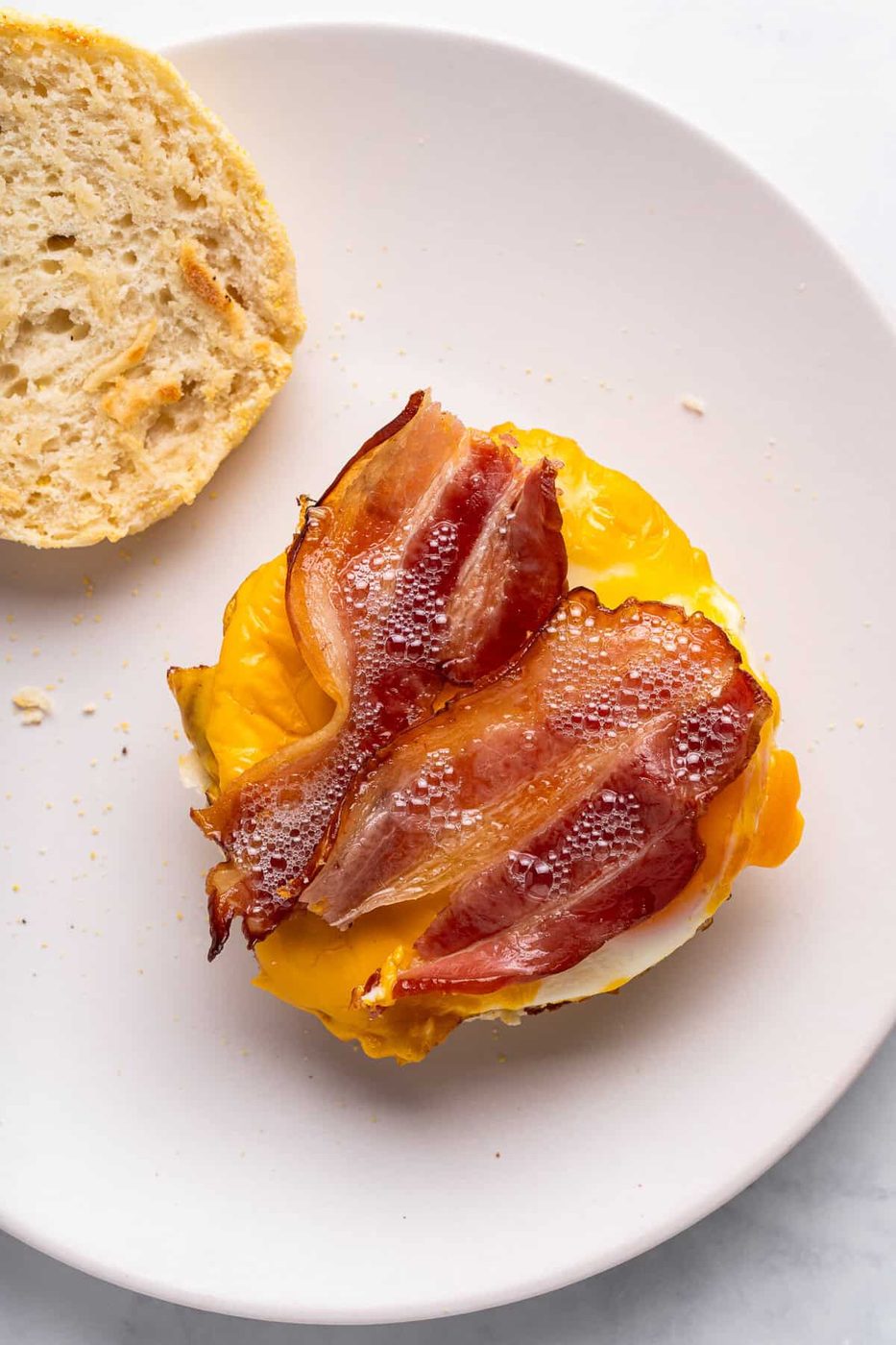 Assembling the homemade egg mcmuffins sandwich with crispy bacon and melted cheese on a fried egg, served on a round white plate with a partially sliced roll on the side.