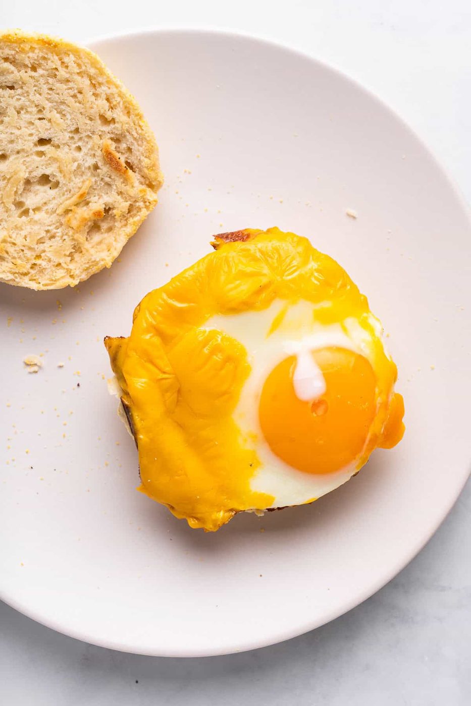 A plate with a toasted English muffin topped with melted cheese and a fried egg with a runny yolk. A slice of bread is on the side. The background is a light, smooth surface.