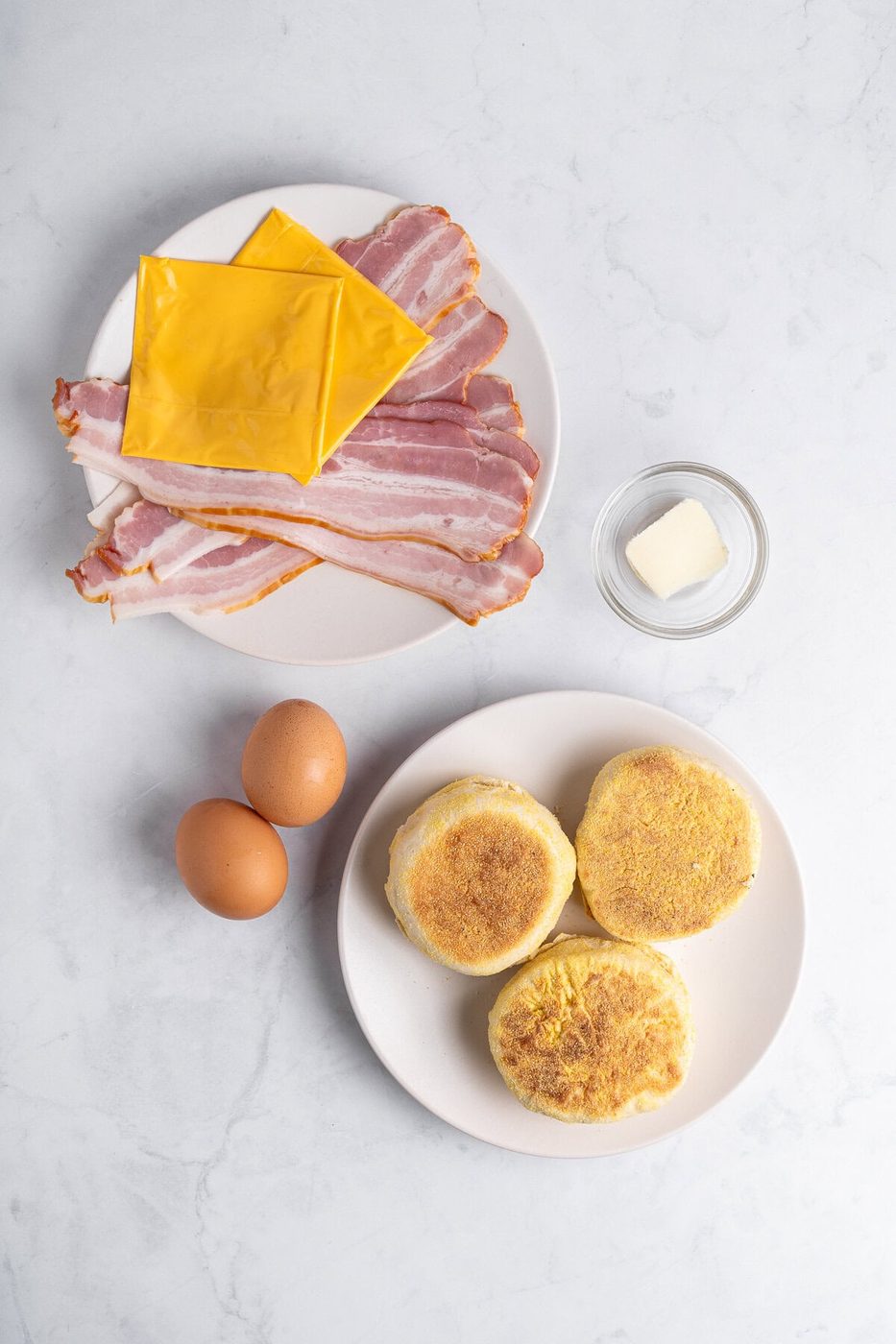 A plate with bacon slices and cheese on a marble surface, alongside a small dish with butter, two eggs, and a plate with two English muffins.