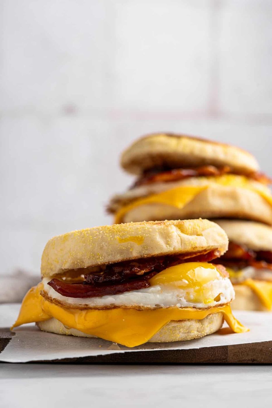 A close-up of homemade egg mcmuffins stacked on top of each other. Each sandwich is made of a toasted English muffin with melted cheese, a fried egg, and crispy bacon. The background is softly blurred.