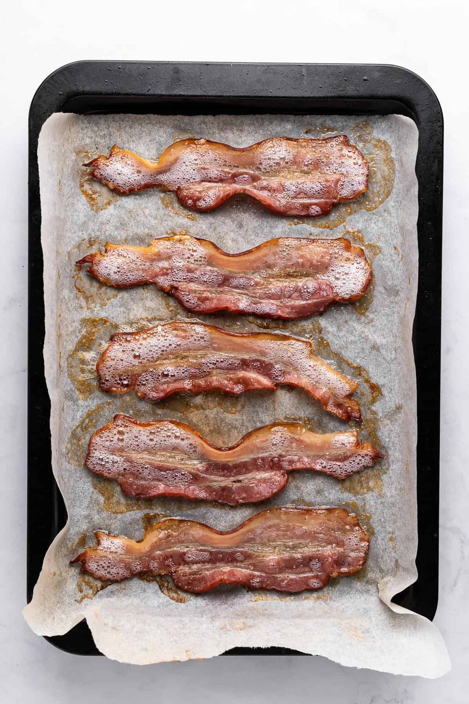 Five slices of cooked bacon on a baking tray lined with parchment paper. The bacon is arranged in a single layer with a crispy appearance. The tray is placed on a light-colored surface.