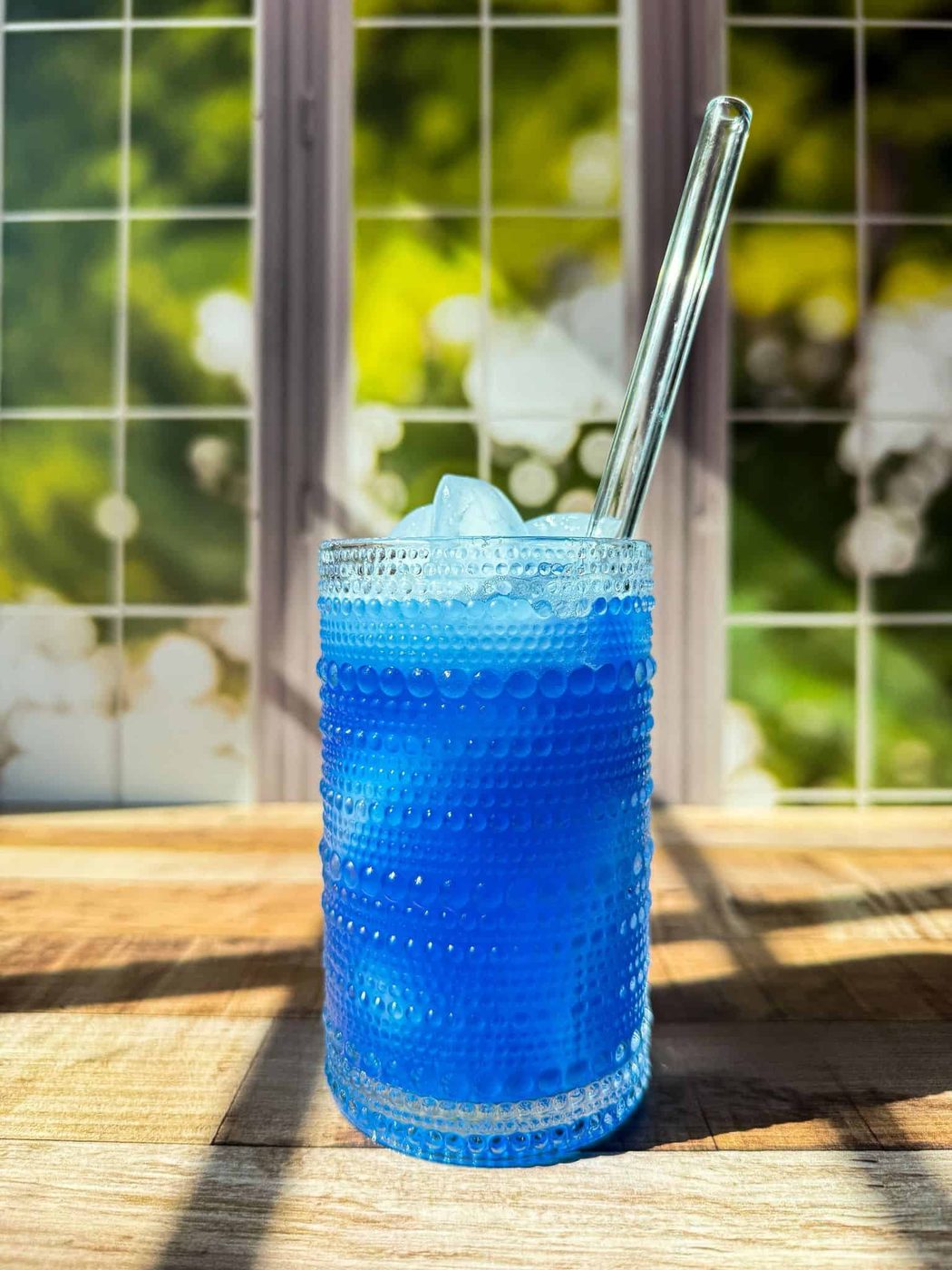 A textured glass filled with homemade gatorade and ice sits on a wooden surface, with a clear straw. Sunlight streams through a window with a green and white background, casting shadows across the scene.