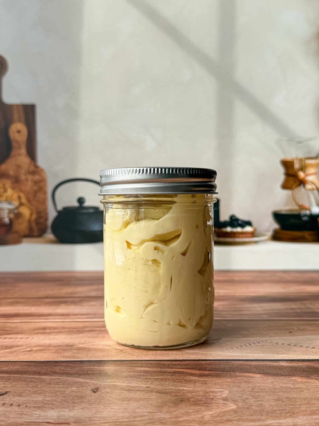 A glass jar with a metal lid filled with creamy yellow homemade mayo sits on a wooden surface in a kitchen, surrounded by blurred utensils and cookware in the background.
