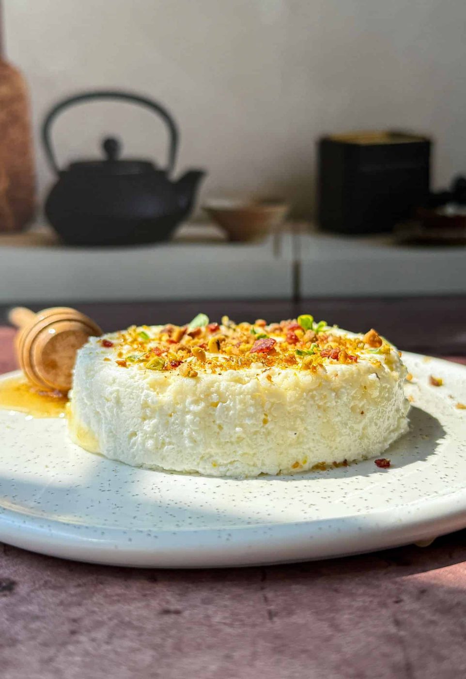 Homemade ricotta topped with chopped nuts and syrup sits on a white plate. A honey dipper with honey rests beside it. The background shows a blurred black teapot and kitchen items.