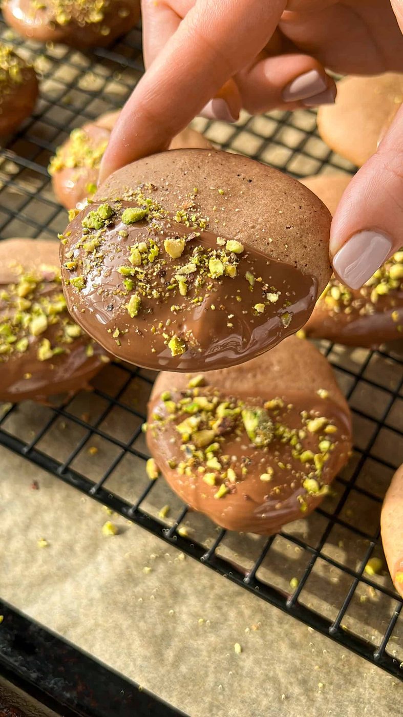 A hand holding a round cookie dipped in chocolate and sprinkled with crushed pistachios, with similar cookies resting on a wire cooling rack in the background.