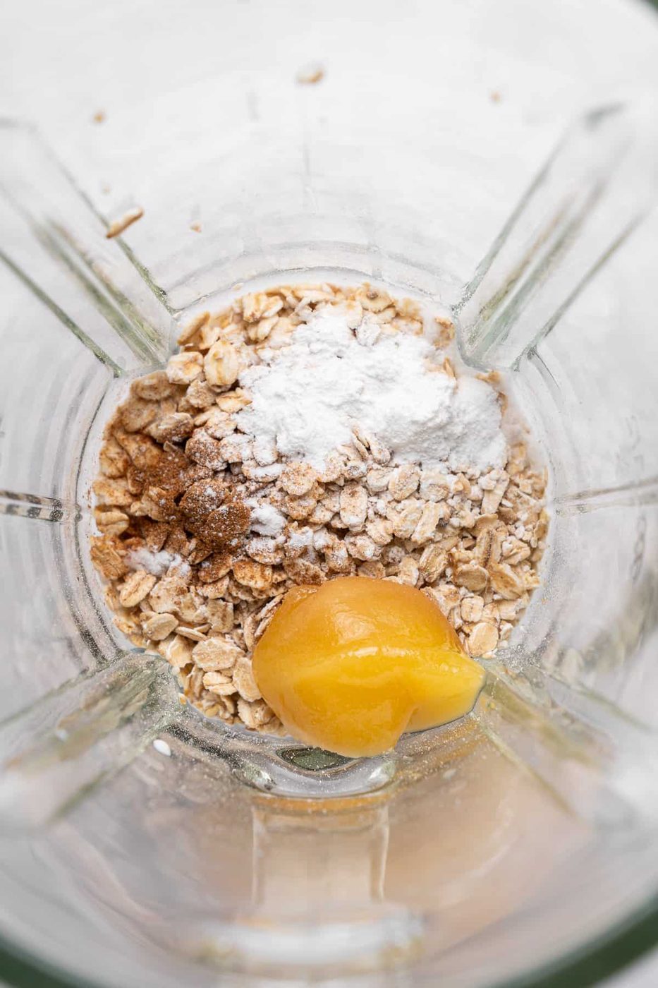 A blender contains oats, a dollop of honey, cinnamon, and a scoop of white powder, likely baking powder, seen from above. The ingredients are ready for blending, suggesting preparation for a smoothie or baking recipe.
