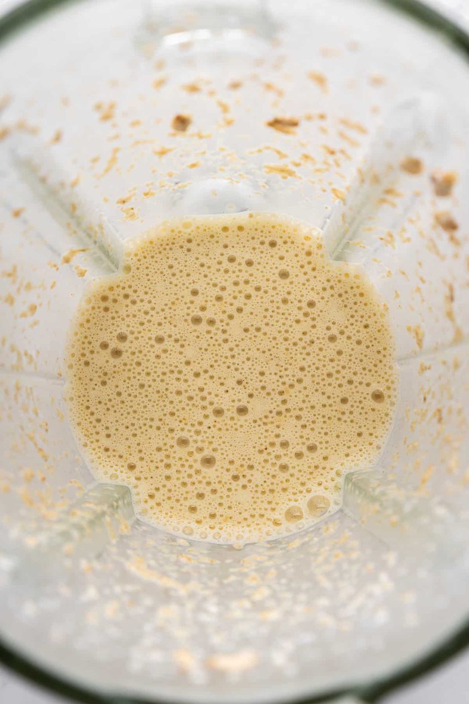 A top view of a blender with a creamy, frothy beige liquid inside, likely a smoothie or shake. The blender shows some light residue on the sides, suggesting recent blending.