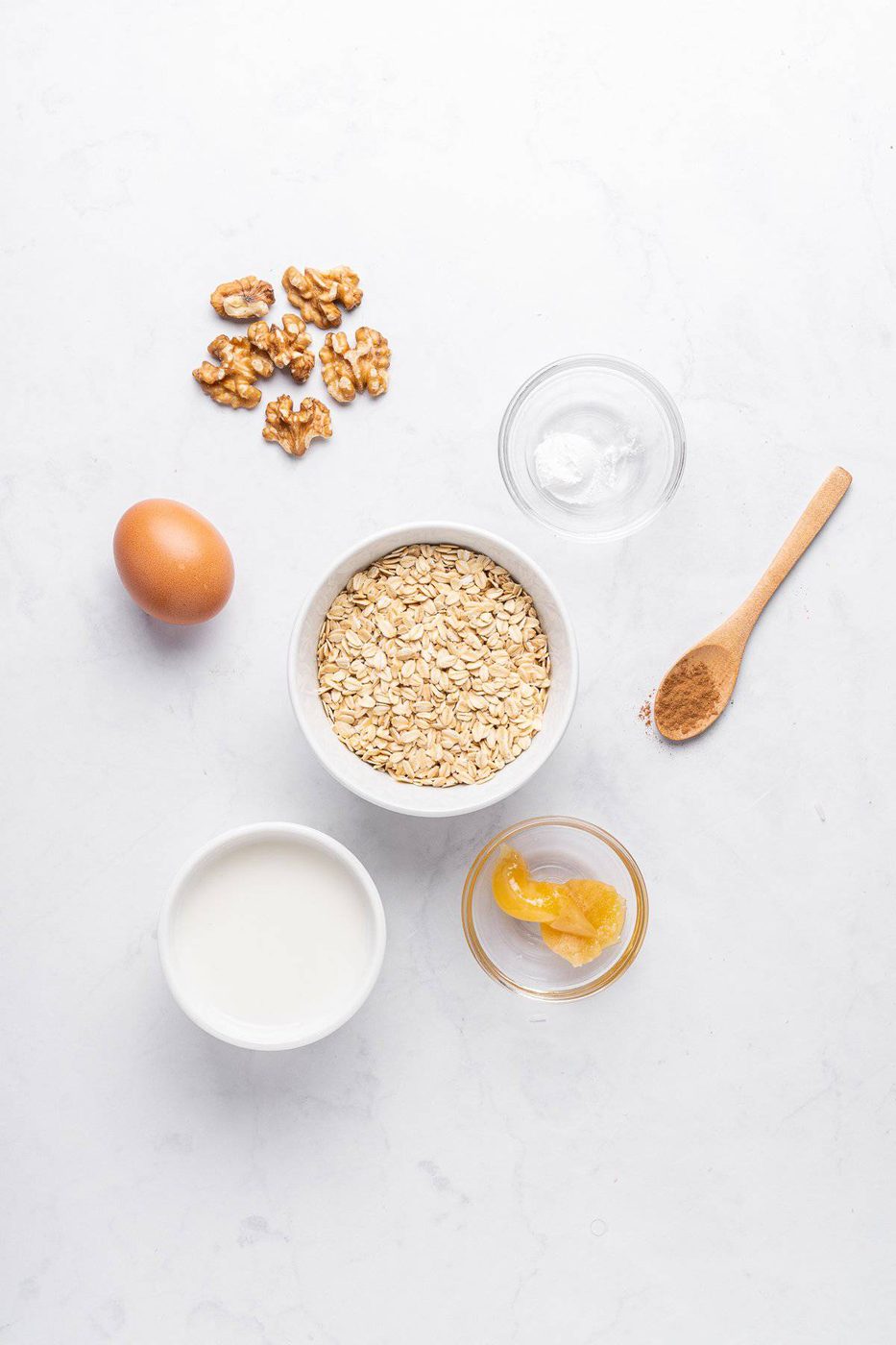 Ingredients for a recipe laid out on a white surface: a bowl of oats, a small bowl of milk, an egg, walnut pieces, a spoonful of cinnamon, a bowl of honey, and a small dish of baking soda.