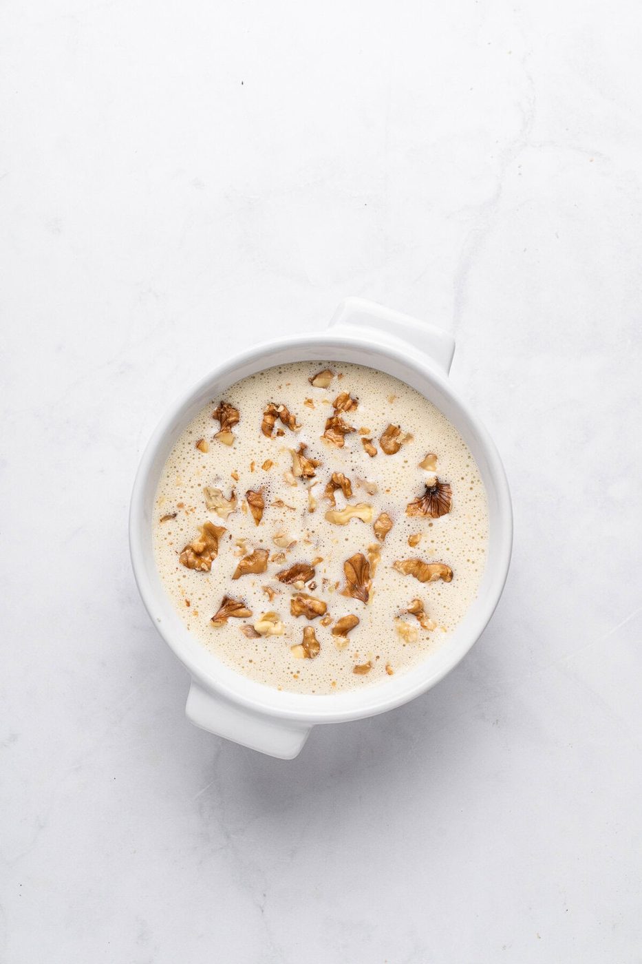 A round white dish filled with creamy pudding, topped with chopped walnuts, sits on a light gray marble surface. The pudding has a smooth texture, and the nuts add a crunchy garnish.