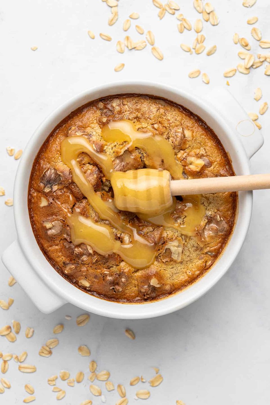 A round baked oatmeal dish in a white ceramic container drizzled with honey, shown on a light surface. Oats and a wooden honey dipper are nearby. The oatmeal has a golden-brown top with visible nuts.