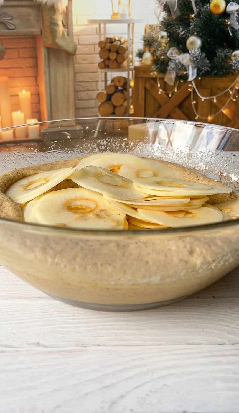 A large glass bowl filled with batter and thinly sliced apples is on a table. In the background, there's a cozy scene with a Christmas tree, a stack of firewood, and candles near a brick fireplace.
